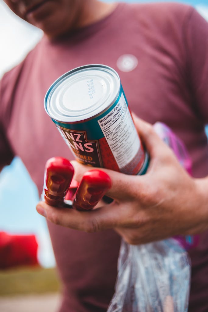 Close-up shot of a man holding a can of beans and tomato paste in an outdoor setting.