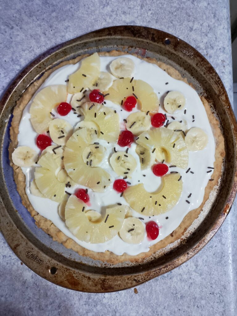 Cookie cased pizza topped with condensed milk sauce and topped with pineapple maraschino cherries and sliced banana