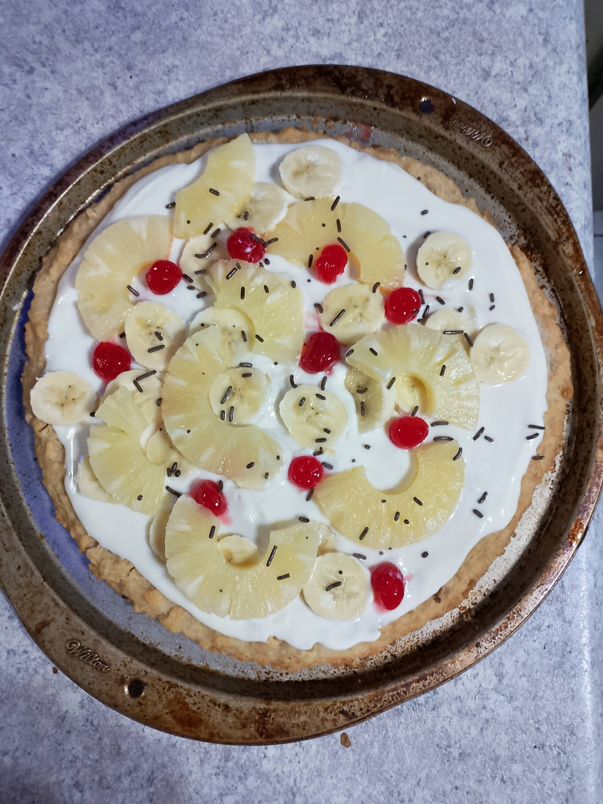 Cookie cased pizza topped with condensed milk sauce and topped with pineapple maraschino cherries and sliced banana